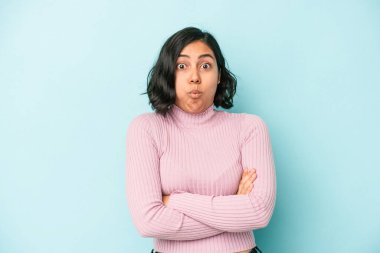 Mavi arka planda izole edilmiş genç Latin kadın yanakları patlatıyor, yorgun bir ifade takınıyor. Yüz ifadesi kavramı.