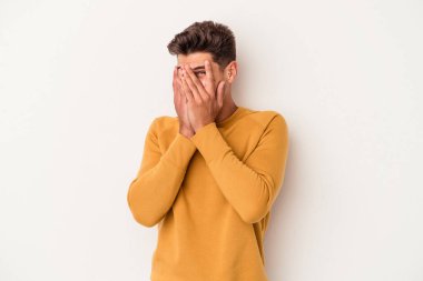 Beyaz arka planda izole edilmiş genç beyaz bir adam kameraya parmaklarıyla göz kırpıyor, yüzünü kapatmaktan utanıyor..