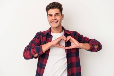 Beyaz arka planda izole edilmiş genç beyaz bir adam gülümsüyor ve elleriyle kalp şekli gösteriyor..