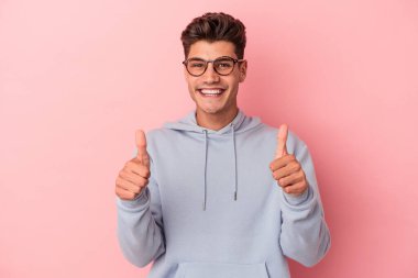 Pembe arka planda izole edilmiş genç beyaz adam gülümsüyor ve başparmağını kaldırıyor.