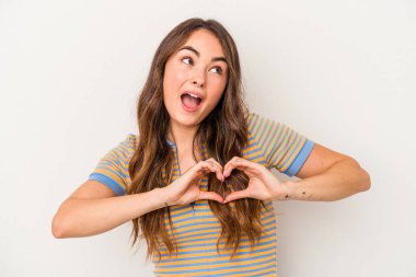 Beyaz arka planda izole edilmiş genç beyaz kadın gülümsüyor ve elleriyle kalp şekli gösteriyor..