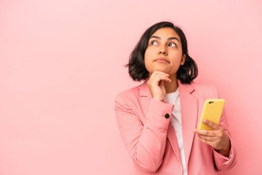 Pembe arka planda cep telefonu tutan genç Latin kadın şüpheli ve şüpheci bir ifadeyle yana bakıyor..