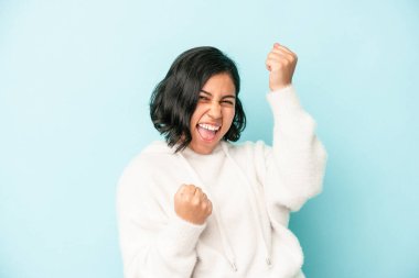 Genç Latin bir kadın mavi arka planda izole edilmiş tasasız ve heyecanlı tezahüratlar yapıyor. Zafer konsepti.