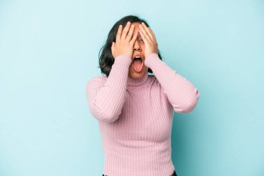 Mavi arka planda izole edilmiş genç Latin kadın yüzünün yarısını avuç içiyle kaplayarak eğleniyor..