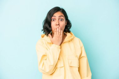 Mavi arka planda izole edilmiş genç Latin kadın şok olmuş, ağzı ellerle kaplanmış, yeni bir şey keşfetmek için sabırsızlanıyor..