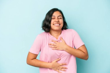 Mavi arka planda yalnız yaşayan genç Latin kadın mutlu bir şekilde gülüyor ve ellerini karnının üstünde tutarak eğleniyor..