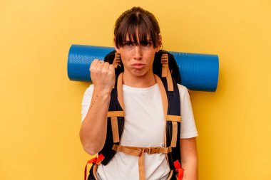 Sarı arka planda izole edilmiş genç Arjantinli kadın kameraya yumruk gösteriyor, agresif yüz ifadesi..