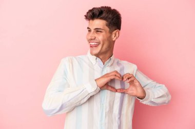 Pembe arka planda izole edilmiş genç beyaz adam gülümsüyor ve elleriyle kalp şeklini gösteriyor..