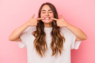 Pembe arka planda izole edilmiş genç beyaz kadın gülümsüyor, parmaklarını ağıza doğrultuyor..