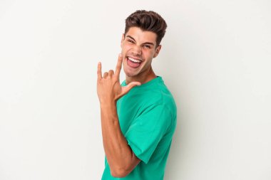 Beyaz arka planda izole edilmiş genç beyaz bir adam parmaklarıyla kaya hareketi gösteriyor.