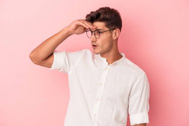 Pembe arka planda izole edilmiş genç beyaz adam uzaklara bakıyor ve elini alnına koyuyor..