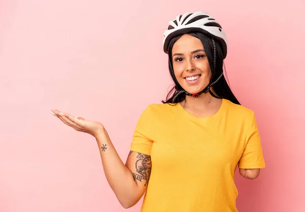 Young caucasian woman with one arm wearing a bike helmet isolated on pink background showing a copy space on a palm and holding another hand on waist.