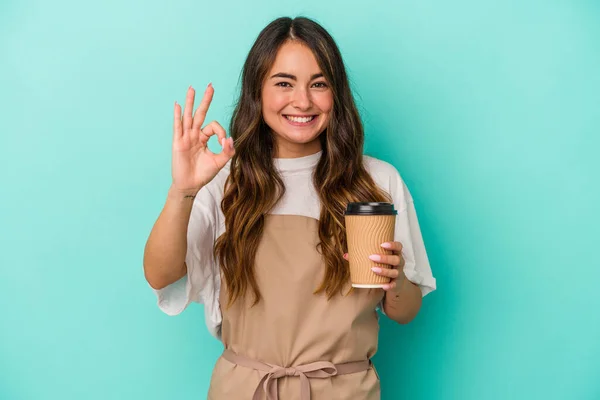 Young caucasian store clerk woman holding a takeaway coffee isolated on blue background cheerful and confident showing ok gesture.