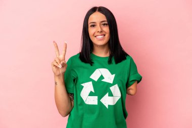 Bir kolu geri dönüştürülmüş beyaz bir kadın. Pembe arka planda izole edilmiş. İki numaranın parmakları olduğunu gösteriyor..