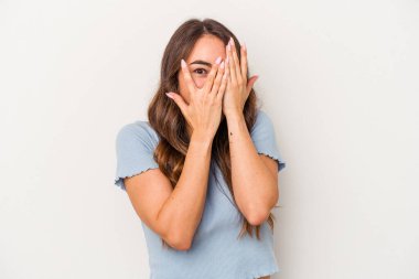 Beyaz arka planda izole edilmiş beyaz tenli genç bir kadın parmaklarının arasından korkmuş ve gergin bir şekilde göz kırpıyor..