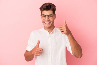 Pembe arka planda izole edilmiş genç beyaz adam iki parmağını da kaldırıyor, gülümsüyor ve kendinden emin..