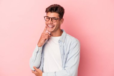 Pembe arka planda izole edilmiş genç beyaz adam gülümsüyor ve kendinden emin, elleriyle çenesine dokunuyor..