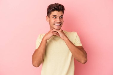 Pembe arka planda izole edilmiş genç beyaz adam ellerini çenesinin altında tutuyor, mutlu bir şekilde yana bakıyor..