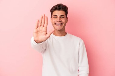 Pembe arka planda izole edilmiş genç beyaz adam gülümseyerek beş numarayı parmaklarıyla gösteriyor..