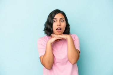 Mavi arka planda izole edilmiş genç Latin bir kadın şans için dua ediyor, şaşırmış ve ağzını açıyor..