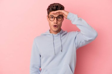 Pembe arka planda izole edilmiş genç beyaz adam uzaklara bakıyor ve elini alnına koyuyor..