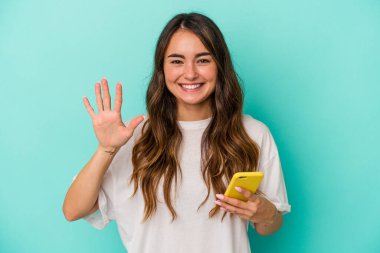 Elinde cep telefonu olan beyaz bir kadın mavi arka planda izole edilmiş. Beş numarayı parmaklarıyla gülümsüyor..