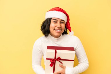 Genç Latin bir kadın Noel 'i kutluyor. Sarı arka planda izole edilmiş bir hediye tutuyor. Gülüyor ve eğleniyor..