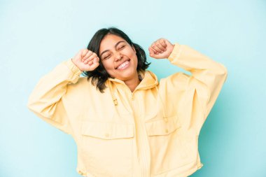 Mavi arka planda izole edilmiş genç Latin kadın özel bir günü kutluyor, atlıyor ve enerjiyle kollarını kaldırıyor..