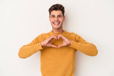 Beyaz arka planda izole edilmiş genç beyaz bir adam gülümsüyor ve elleriyle kalp şekli gösteriyor..