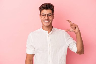 Pembe arka planda izole edilmiş genç beyaz adam işaret parmaklarıyla küçük bir şey tutuyor, gülümsüyor ve kendinden emin..