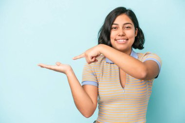 Mavi arka planda izole edilmiş genç Latin bir kadın, avucunun içinde bir fotokopi alanı tutarken heyecanlı..