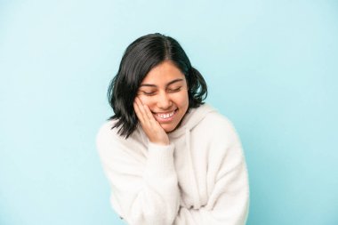 Mavi arka planda yalnız yaşayan genç Latin kadın mutlu bir şekilde gülüyor ve ellerini karnının üstünde tutarak eğleniyor..