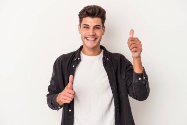 Beyaz arka planda izole edilmiş genç beyaz bir adam iki parmağını da kaldırıyor, gülümsüyor ve kendinden emin..