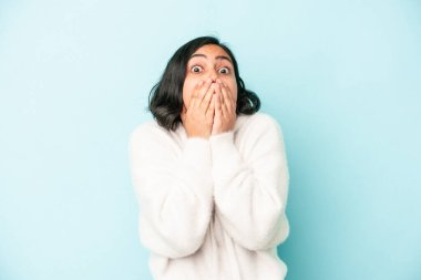 Mavi arka planda izole edilmiş genç Latin kadın şok olmuş, ağzı ellerle kaplanmış, yeni bir şey keşfetmek için sabırsızlanıyor..