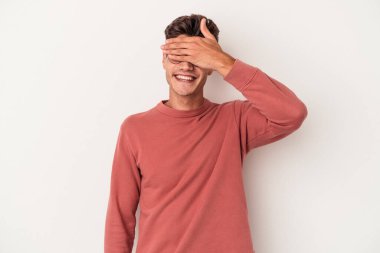 Beyaz arka planda soyutlanmış beyaz tenli genç bir adam elleriyle gözlerini kapatıyor, geniş bir gülümsemeyle bir sürpriz bekliyor..