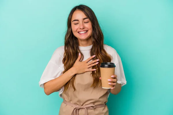 Young caucasian store clerk woman holding a takeaway coffee isolated on blue background laughs out loudly keeping hand on chest.