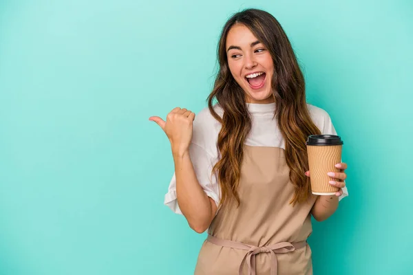 Young caucasian store clerk woman holding a takeaway coffee isolated on blue background points with thumb finger away, laughing and carefree.