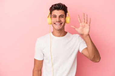 Pembe arka planda izole edilmiş müzik dinleyen beyaz bir adam gülümseyerek beş numarayı parmaklarıyla gösteriyor..