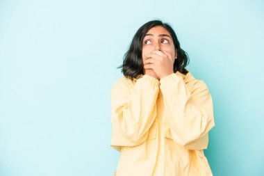 Genç Latin bir kadın mavi arka planda izole edilmiş. Düşünceli bir şekilde bir fotokopi odasına bakıyor. Ağzını eliyle kapatıyor..