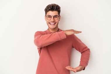 Beyaz arka planda izole edilmiş genç beyaz bir adam işaret parmaklarıyla küçük bir şey tutuyor, gülümsüyor ve kendinden emin..