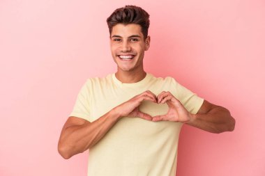 Pembe arka planda izole edilmiş genç beyaz adam gülümsüyor ve elleriyle kalp şeklini gösteriyor..