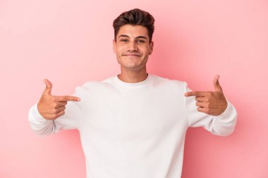 Pembe arka planda izole edilmiş genç beyaz bir adam gömlek fotokopi alanını elle gösteriyor, gururlu ve kendinden emin.