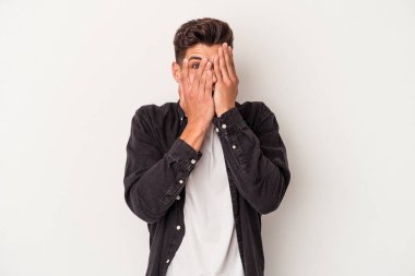 Beyaz arka planda izole edilmiş genç beyaz bir adam parmaklarının arasından korkmuş ve gergin bir şekilde göz kırpıyor..