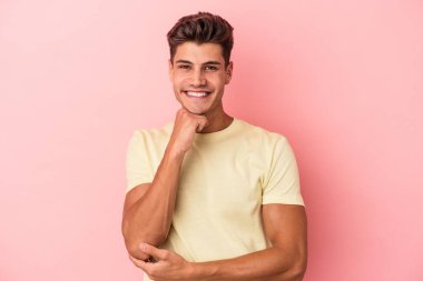 Pembe arka planda izole edilmiş genç beyaz adam gülümsüyor ve kendinden emin, elleriyle çenesine dokunuyor..