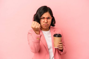 Elinde kahve tutan genç Latin kadın pembe arka planda izole edilmiş yumruğunu kameraya gösteren agresif yüz ifadesi..