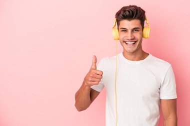 Pembe arka planda izole edilmiş müzik dinleyen beyaz bir adam gülümsüyor ve başparmağını kaldırıyor.