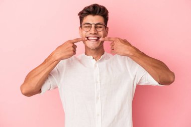 Pembe arka planda izole edilmiş genç beyaz adam gülümsüyor, parmaklarını ağıza doğrultuyor..