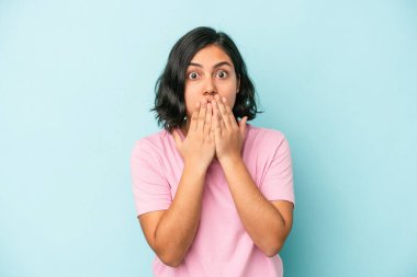 Mavi arka planda izole edilmiş genç Latin kadın şok olmuş, ağzı ellerle kaplanmış, yeni bir şey keşfetmek için sabırsızlanıyor..
