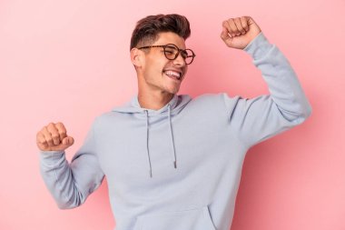 Pembe arka planda soyutlanmış genç beyaz adam özel bir günü kutluyor, atlıyor ve enerjiyle kollarını kaldırıyor..