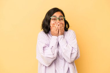 Sarı arka planda izole edilmiş genç Latin bir kadın bir şeye gülüyor, elleriyle ağzını kapatıyor..